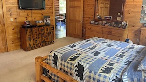 Quarto com painéis de madeira, uma cama, televisão fixada na parede e cômoda com fotografias de família.