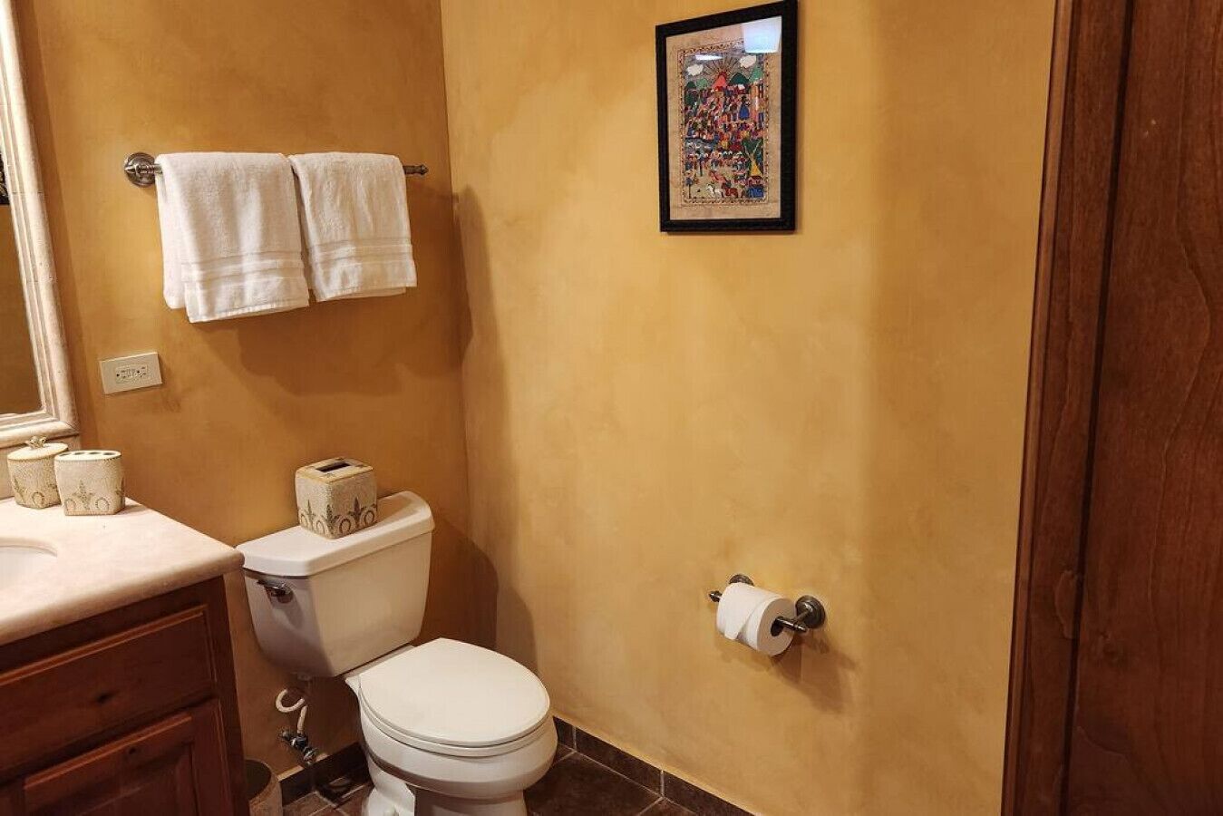 Ein Badezimmer mit Toilette, zwei weiße Handtücher hängen an einem Handtuchhalter, einem gerahmten Bild an der Wand und einer Toilettenpapierrolle.