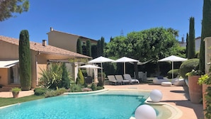 A swimming pool with lounge chairs and umbrellas, surrounded by well-maintained landscaping and a building with a tiled roof.