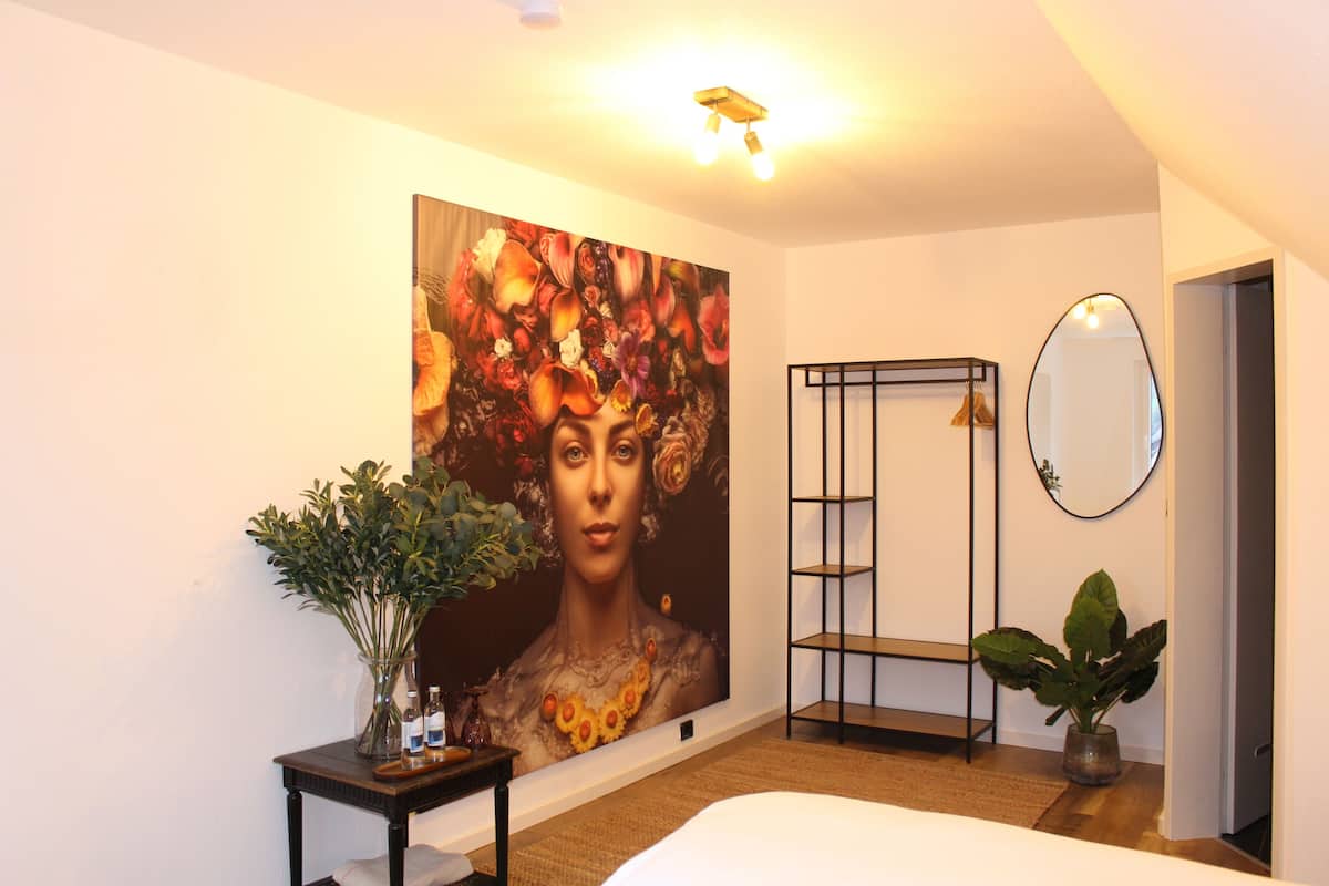 A bedroom with a large floral painting, a bedside table with a plant, a mirror, and a wardrobe.