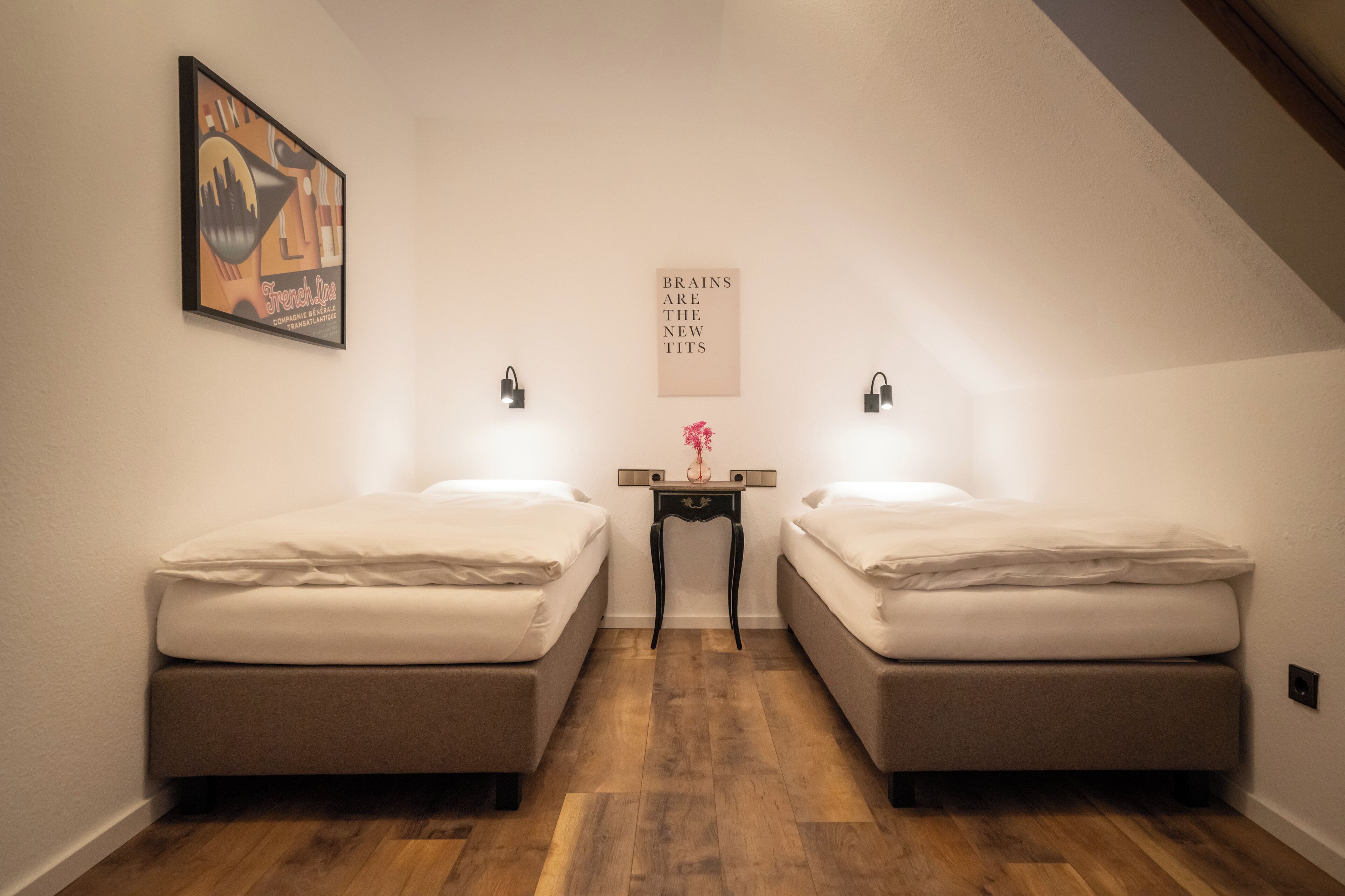 Um quarto pequeno e acolhedor com duas camas de solteiro, uma mesinha e um quadro na parede.