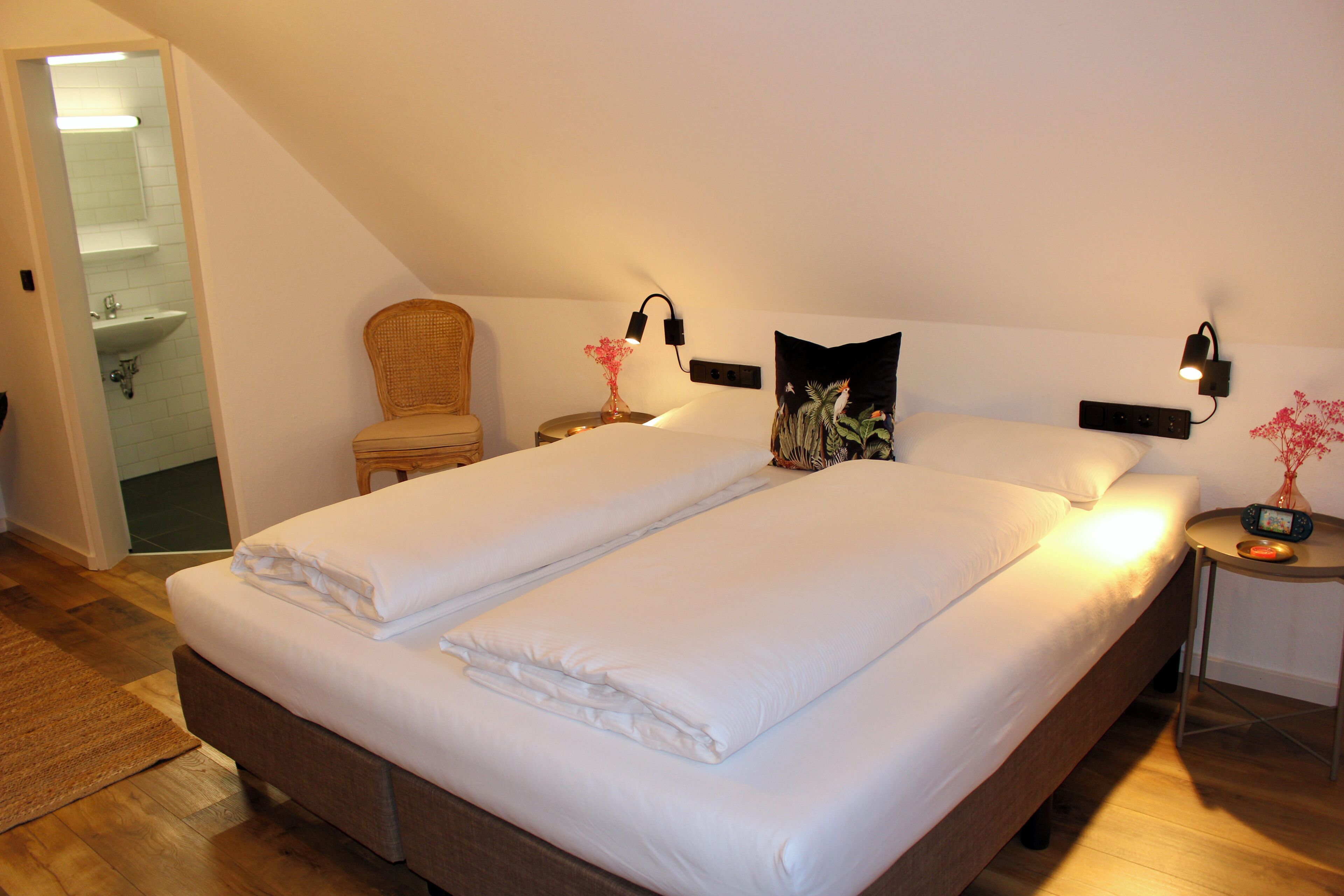 Un lit double avec du linge de lit blanc, une table de chevet avec une lampe, une chaise et une petite table sur laquelle est posé un vase de fleurs.
