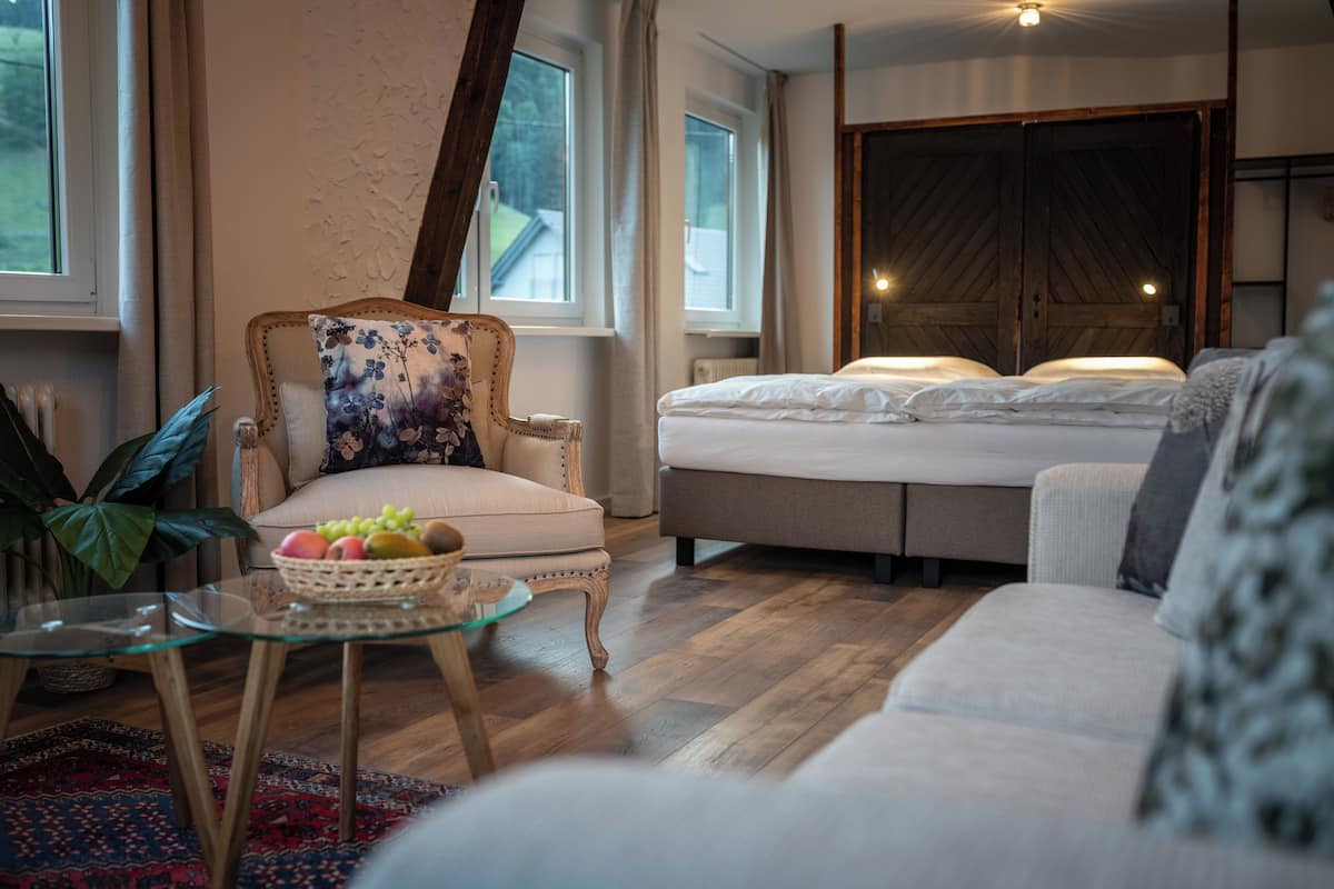 A cozy room with a bed, a sofa, a chair with a floral cushion, and a glass table with a fruit basket.