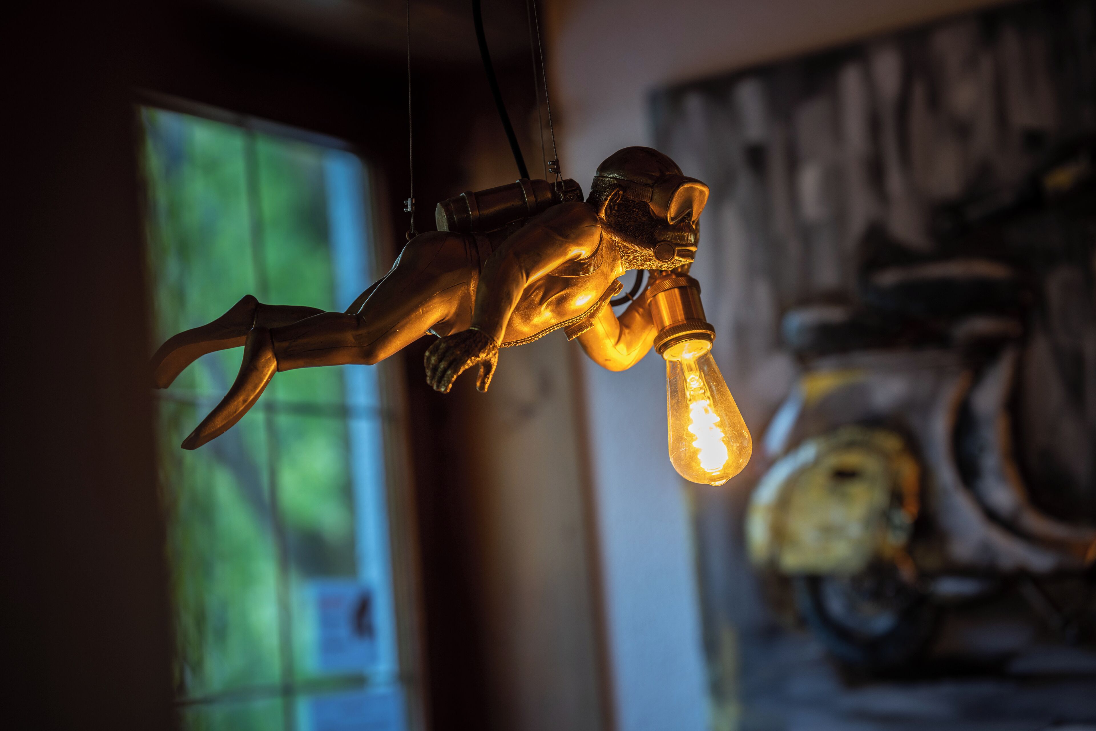A sculpture of a figure with a light bulb for a head, suspended in a room with a blurred background.