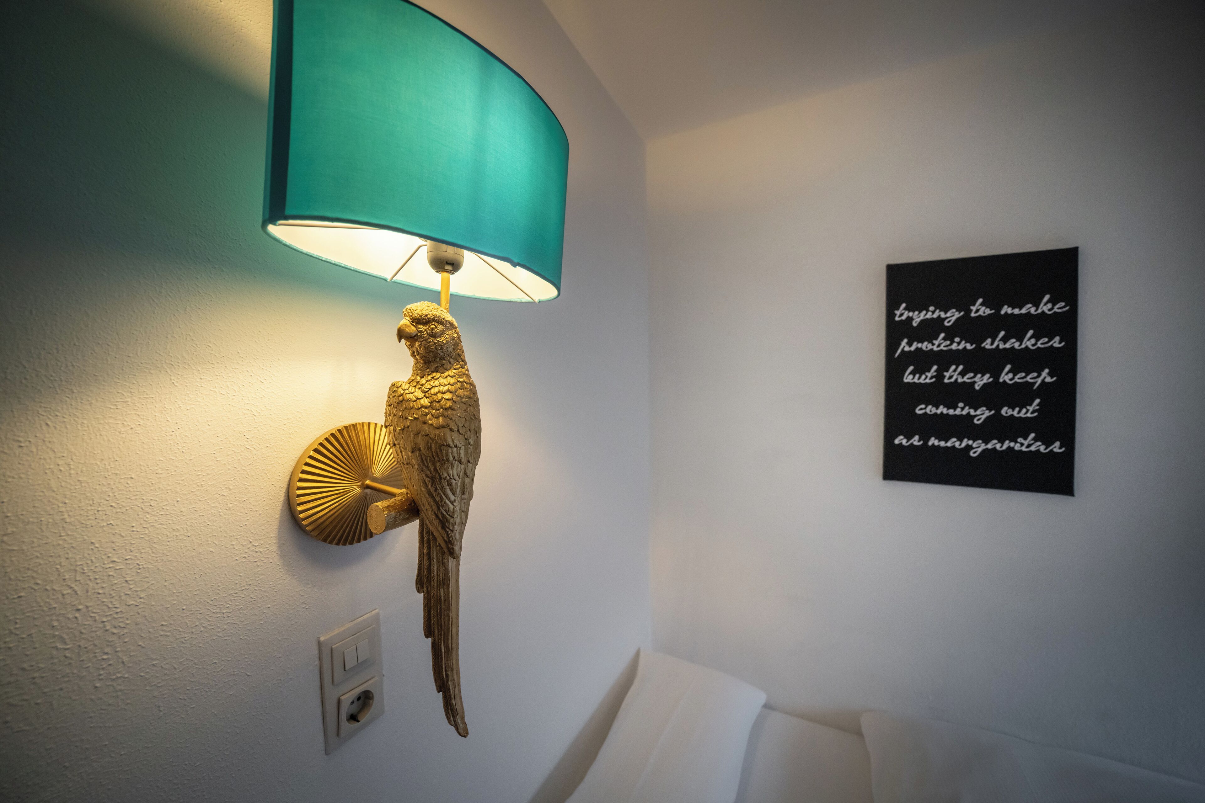 Una lampada a parete con un paralume color petrolio e un motivo ad uccello dorato, una parete bianca e un cartello nero con scritte bianche.