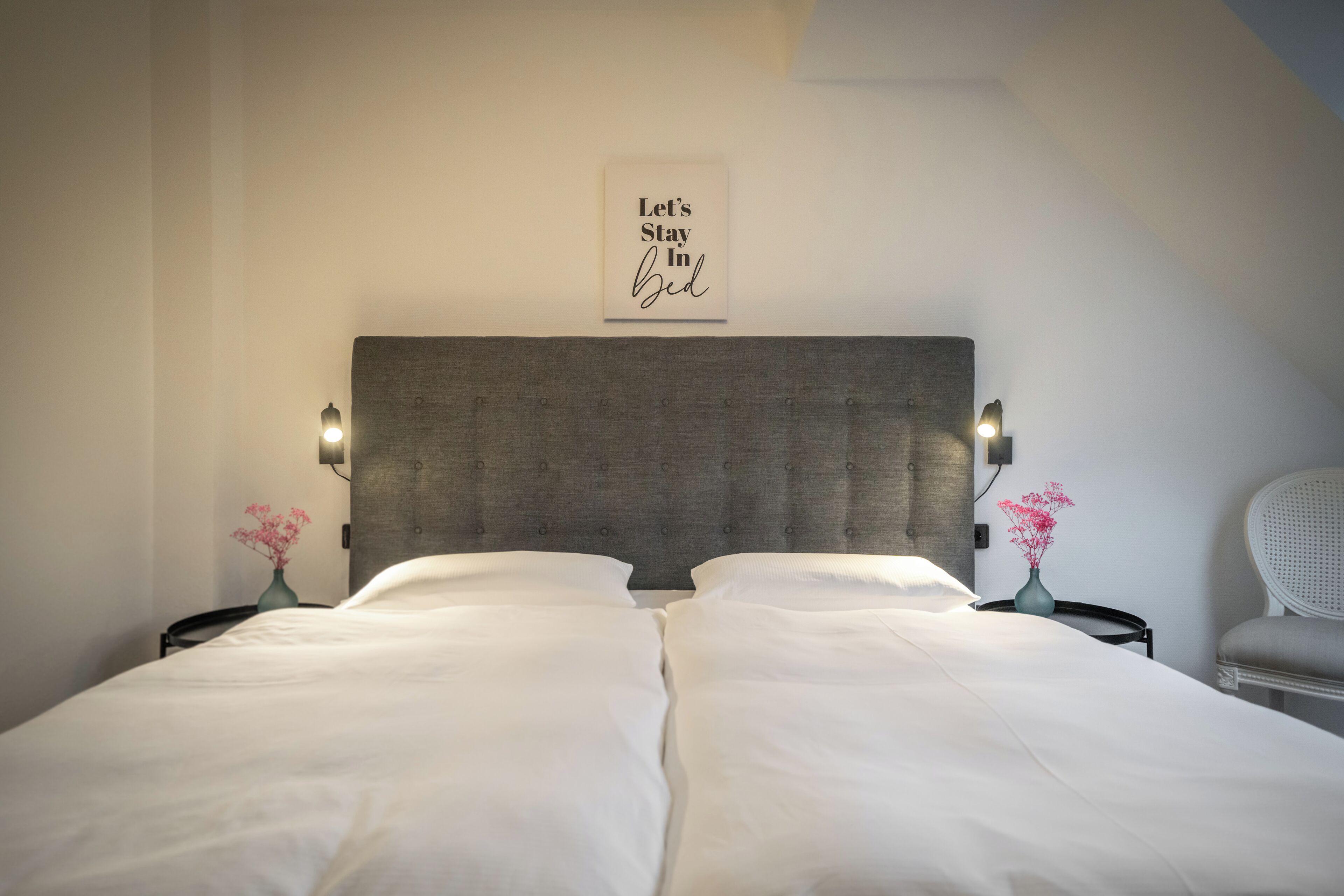 A neatly made twin bed with a grey upholstered headboard, two bedside tables with lamps, and a wall-mounted sign that reads "Let's Stay In Bed".
