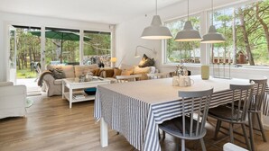 A modern living room with a large window, a dining area with striped tablecloths, and a cozy seating area with a sofa and cushions.
