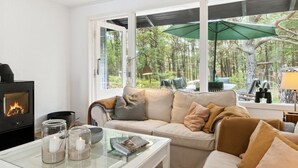 A cozy living room with a fireplace, a sofa, a coffee table with candles, and a view of a forest through large windows.