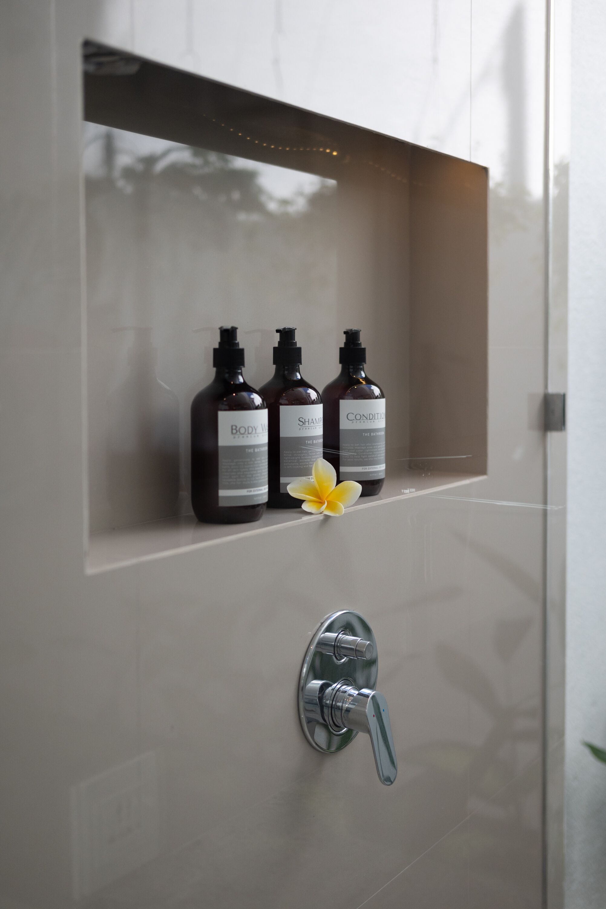 A bathroom with a shower, a shelf with three bottles of body care products, and a yellow flower.