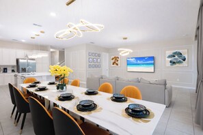 Una cucina moderna con mobili bianchi, un'area pranzo con tavolo bianco apparecchiato per sei persone e un divano grigio nella zona living.
