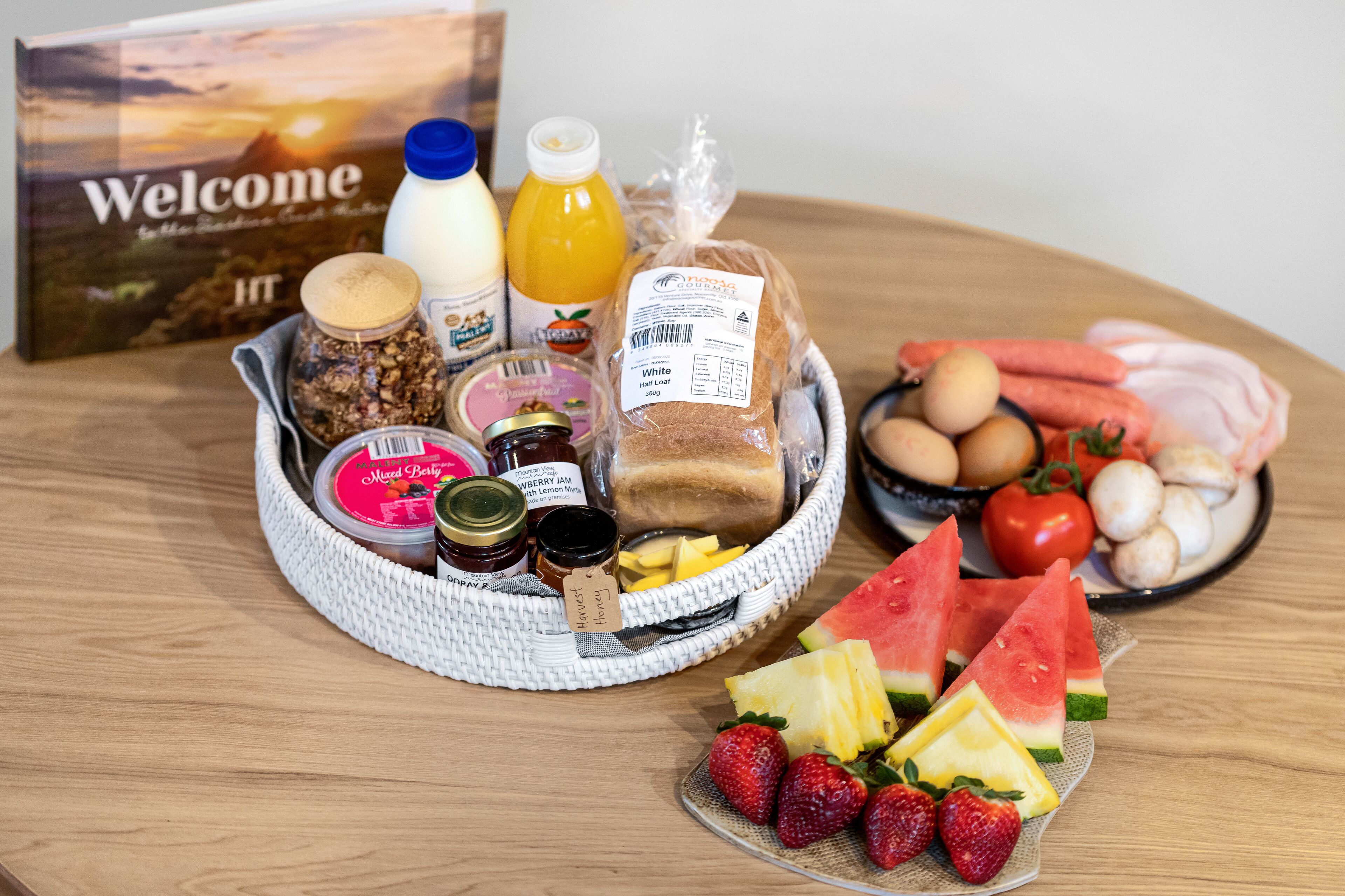 Una mesa de madera con un desayuno que incluye pan, huevos, tomates, sandía, fresas y diferentes condimentos.