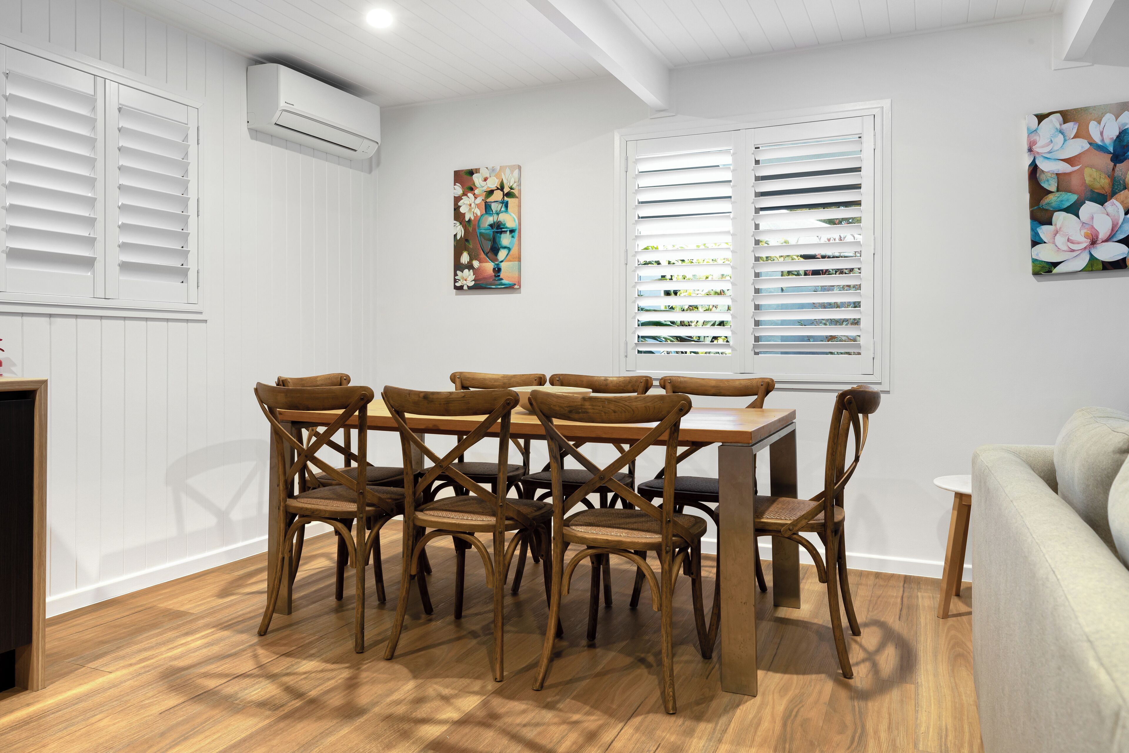 A dining area with a wooden table and chairs, white walls, and two windows with blinds.