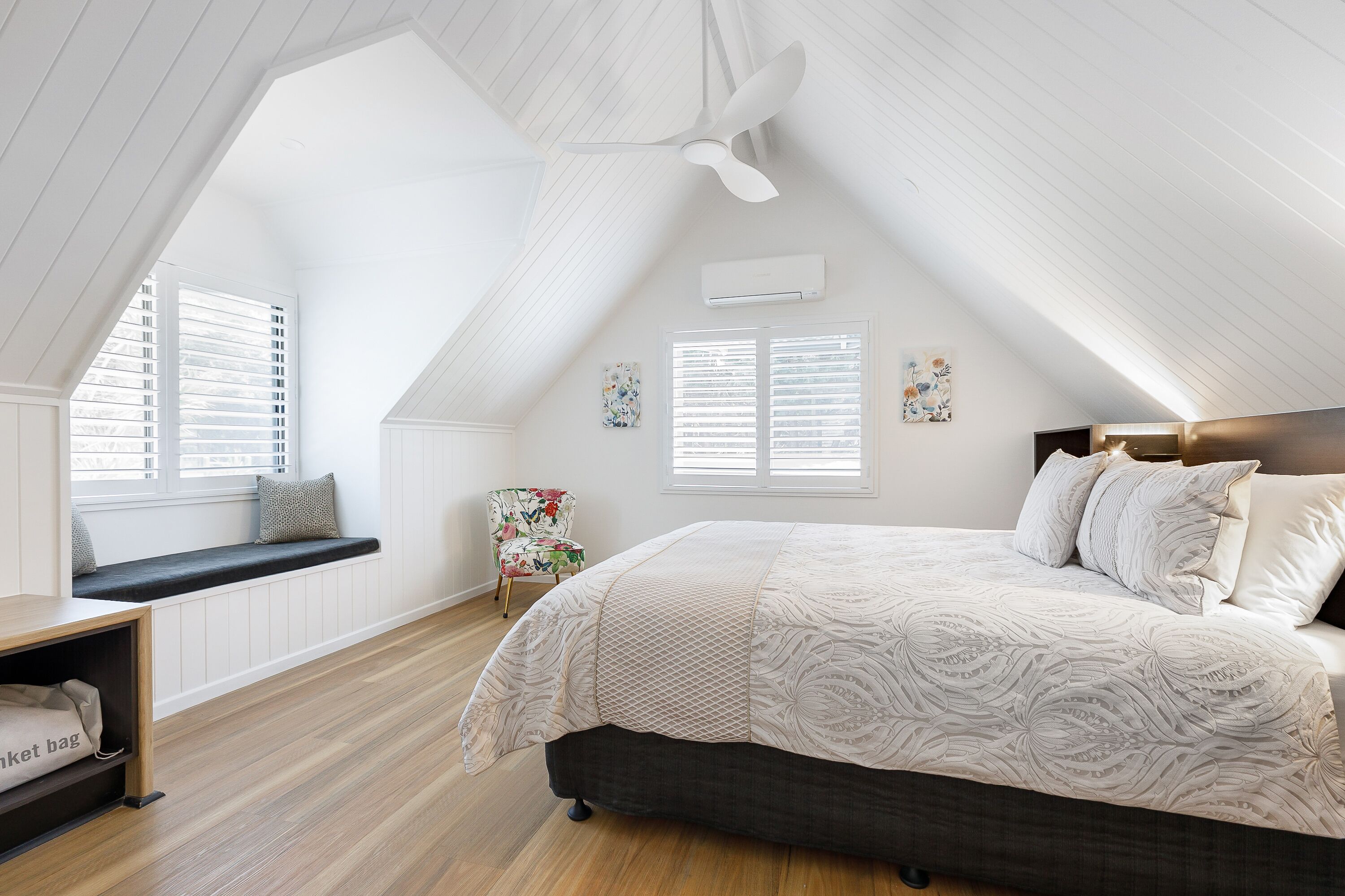 A bedroom with a sloped ceiling, a bed with a patterned bedspread, a chair, and a bench by the window.