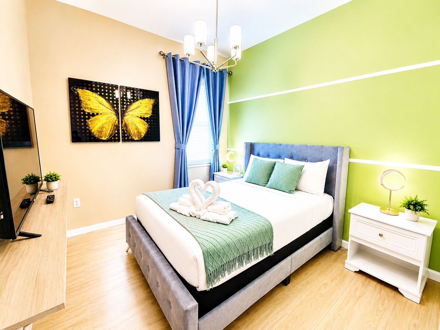 Een moderne slaapkamer met een bed, een nachtkastje, een spiegel, een aan de muur bevestigde televisie en een schilderij van een vlinder.