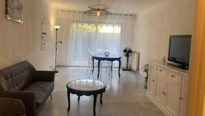 A living room with a sofa, a round table, a TV on a cabinet, and a dining area with chairs and a table. - Grand T3 Terrasses Jardin (Antibes)