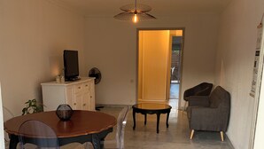 A dining area with a marble floor, a wooden table, and chairs. There is a television on a cabinet, a plant, and a ceiling fan. - Grand T3 Terrasses Jardin (Antibes)