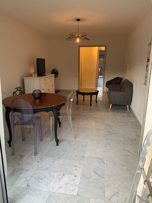 Uma sala de jantar com piso de mármore, mesa de madeira e cadeiras. Há uma televisão em um armário, uma planta e um ventilador de teto.