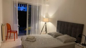 A bedroom with a bed, a nightstand, a lamp, a chair, and a view of the outside. - Grand T3 Terrasses Jardin (Antibes)