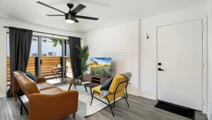 A modern living room with a sofa, armchair, and a TV on a stand. There is a ceiling fan, a door, and a window with curtains. - King Bed, Parking, Pool, Museums, Aquarium! (Tempe)