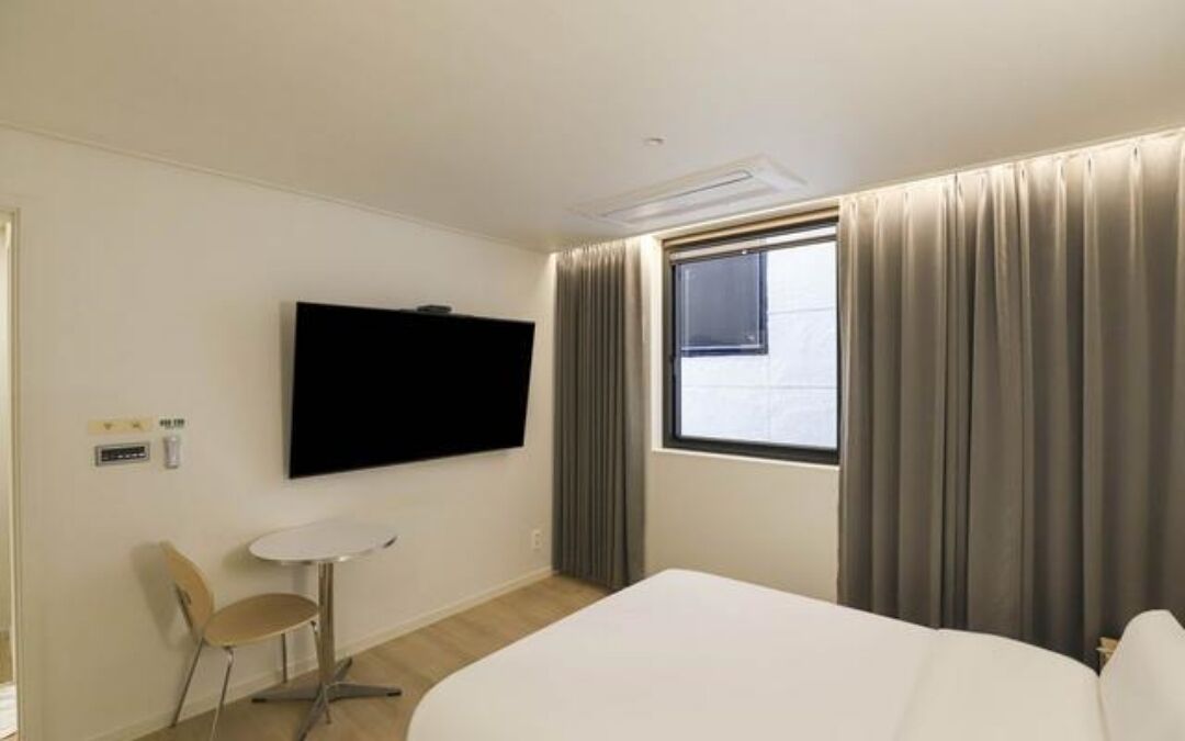 A hotel room with a bed, a TV mounted on the wall, a small table with a chair, and a window with curtains.