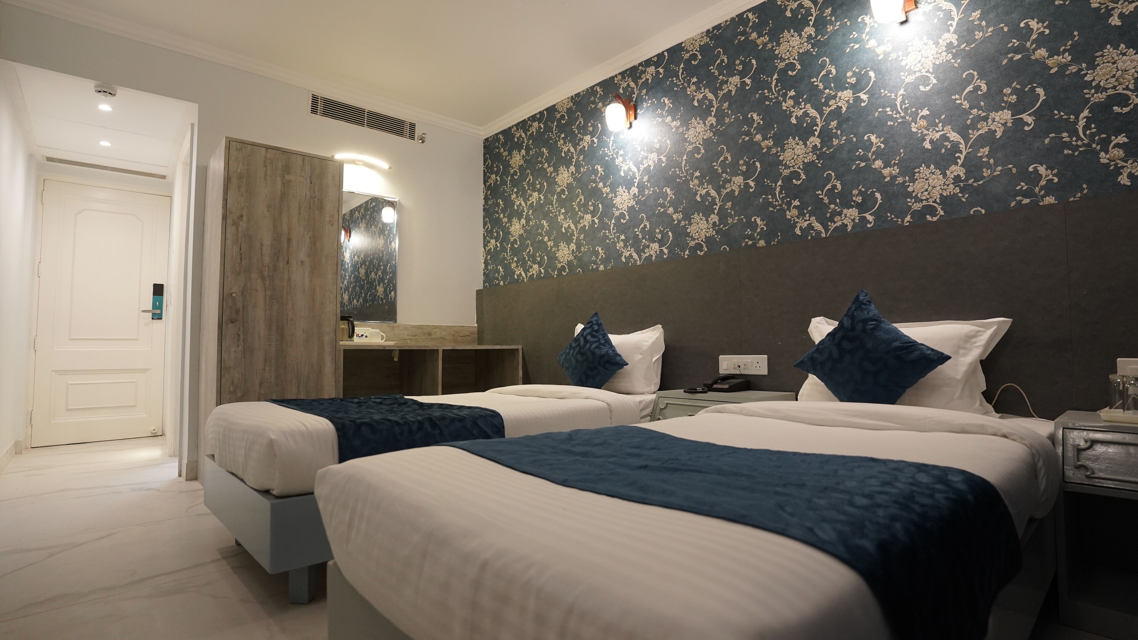 A hotel room with two beds, a wooden wardrobe, a mirror, a nightstand, and a wall with a floral pattern.