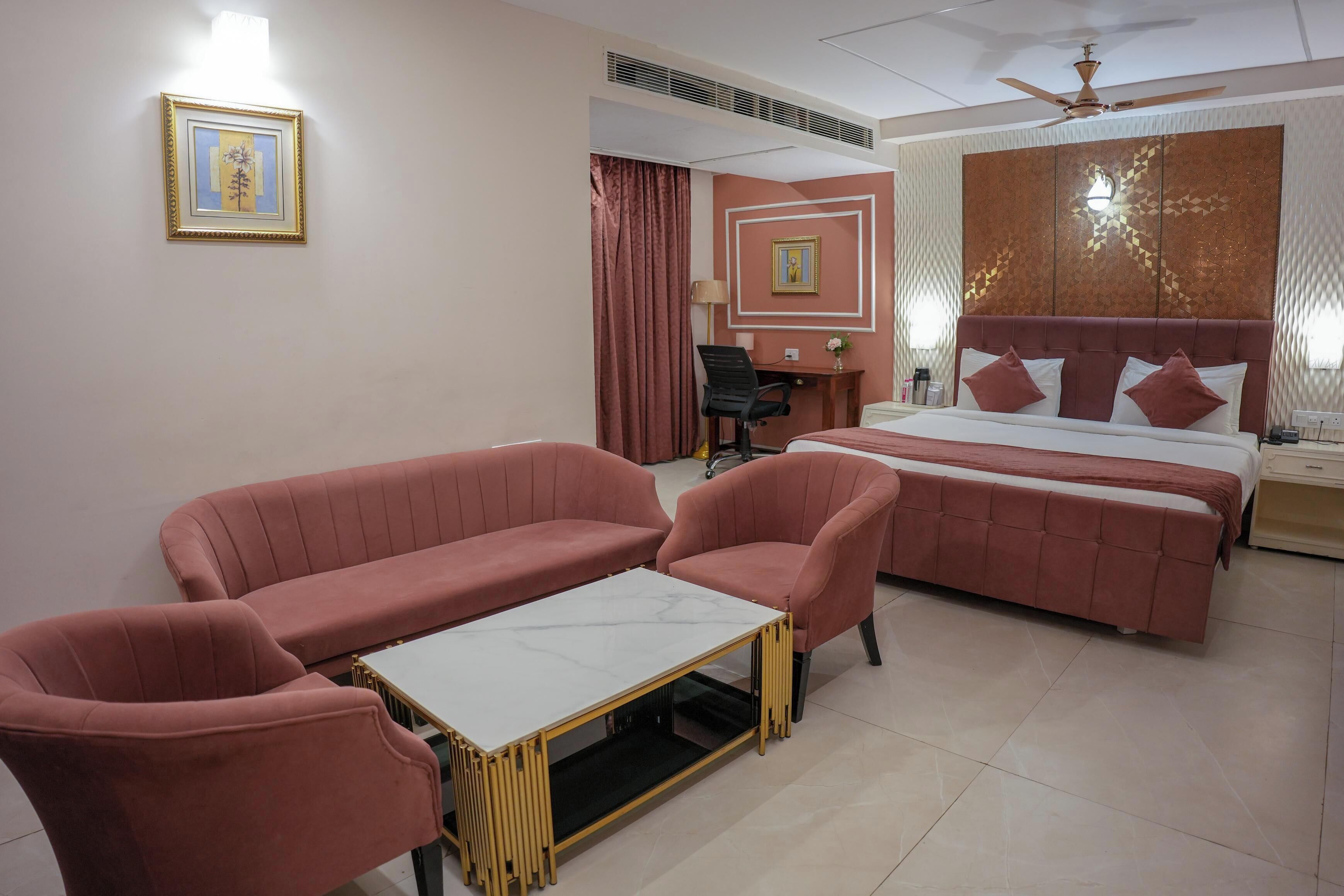 Habitación de hotel con cama, dos sillones rosa, una mesita, un escritorio y un ventilador de techo.