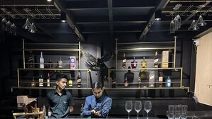 Un espace bar avec deux barmen, un comptoir en marbre, des étagères remplies de bouteilles et des luminaires suspendus.