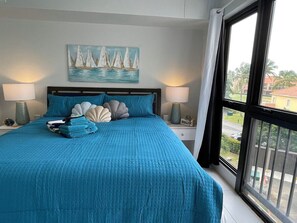 Une chambre à coucher avec un grand lit, deux lampes de chevet, un tableau représentant des voiliers et une vue sur l’extérieur.