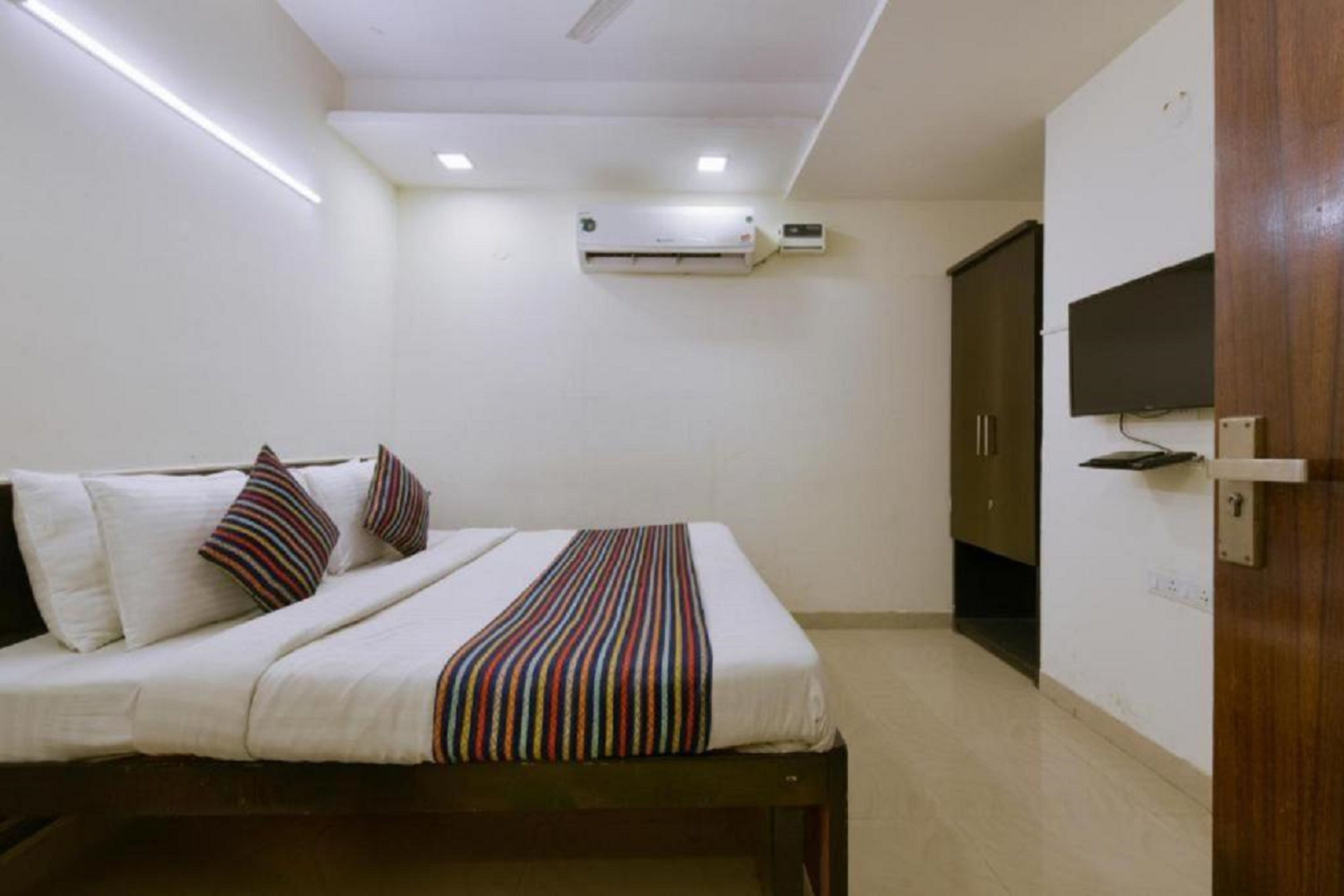 A bedroom with a bed, pillows, a striped blanket, an air conditioning unit, a television, and a wardrobe.