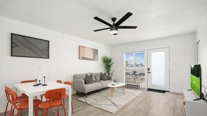 A modern living room with a white sofa, orange chairs, a dining table with candles, a ceiling fan, and a TV. - King Bed, Pool, Parking, Theme Park, Gym SOON! (Phoenix)