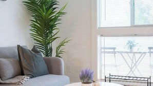 A living room with a grey sofa, a large green plant, a small table with a vase of purple flowers, and a window with blinds. - King Bed, Pool, Parking, Theme Park, Gym SOON! (Phoenix)