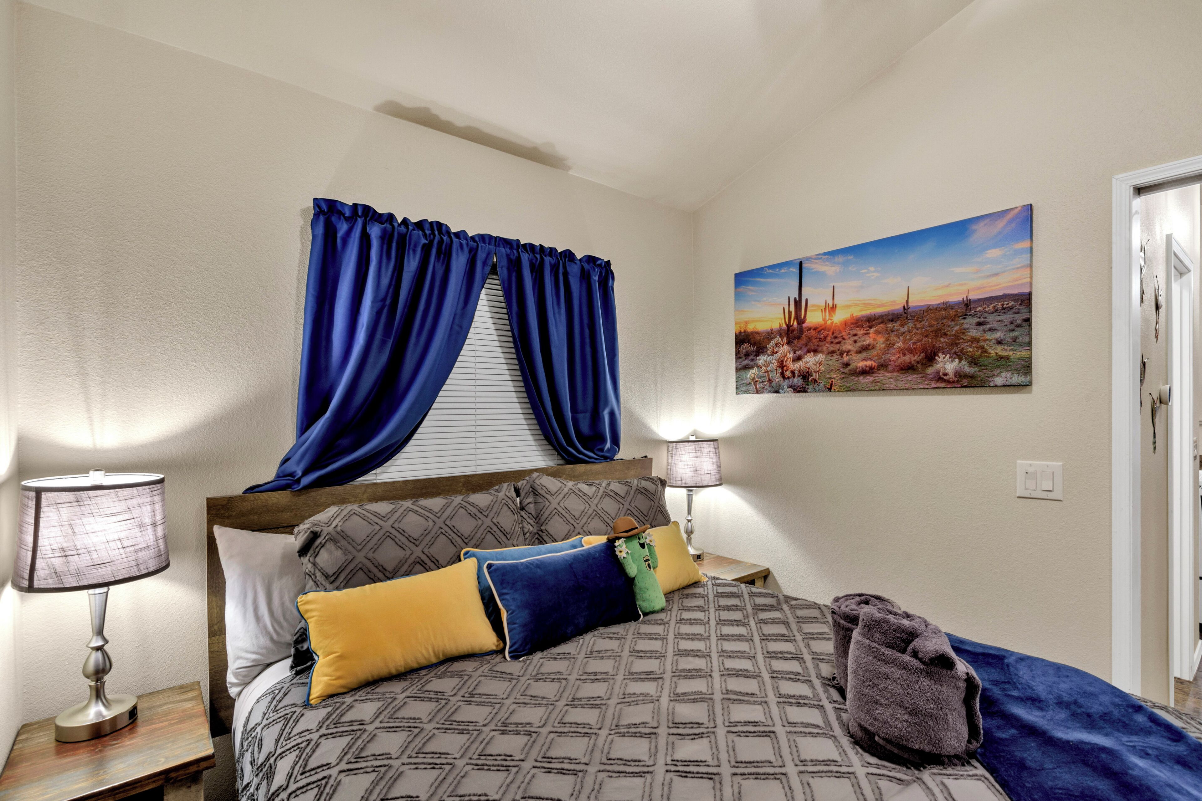 Un dormitorio con cama, almohadas, una mesita de noche con lámpara, una ventana con cortinas y un cuadro en la pared.