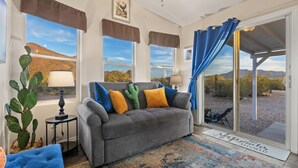 A living room with a grey sofa, blue and yellow cushions, a cactus plant, and a view of the desert through large windows. - Saguaro Sunset Oasis (Tucson)