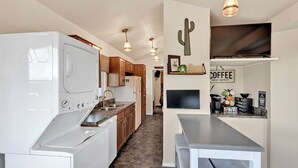 A compact kitchen with a built-in washer and dryer, wooden cabinets, a countertop, and a flat-screen TV mounted above a coffee station. - Saguaro Sunset Oasis (Tucson)