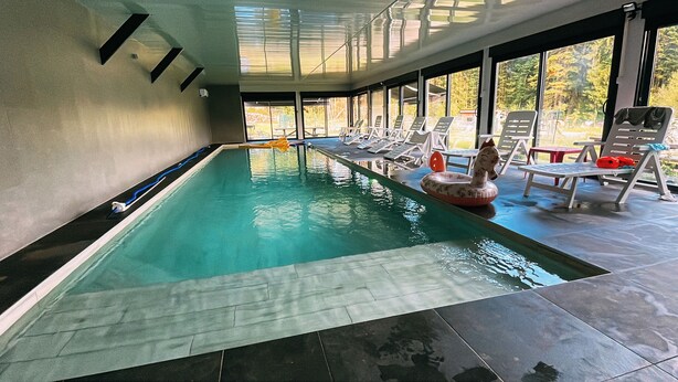 Una piscina interior con agua cristalina, rodeada de sillones de descanso y una zona cubierta con ventanales grandes.