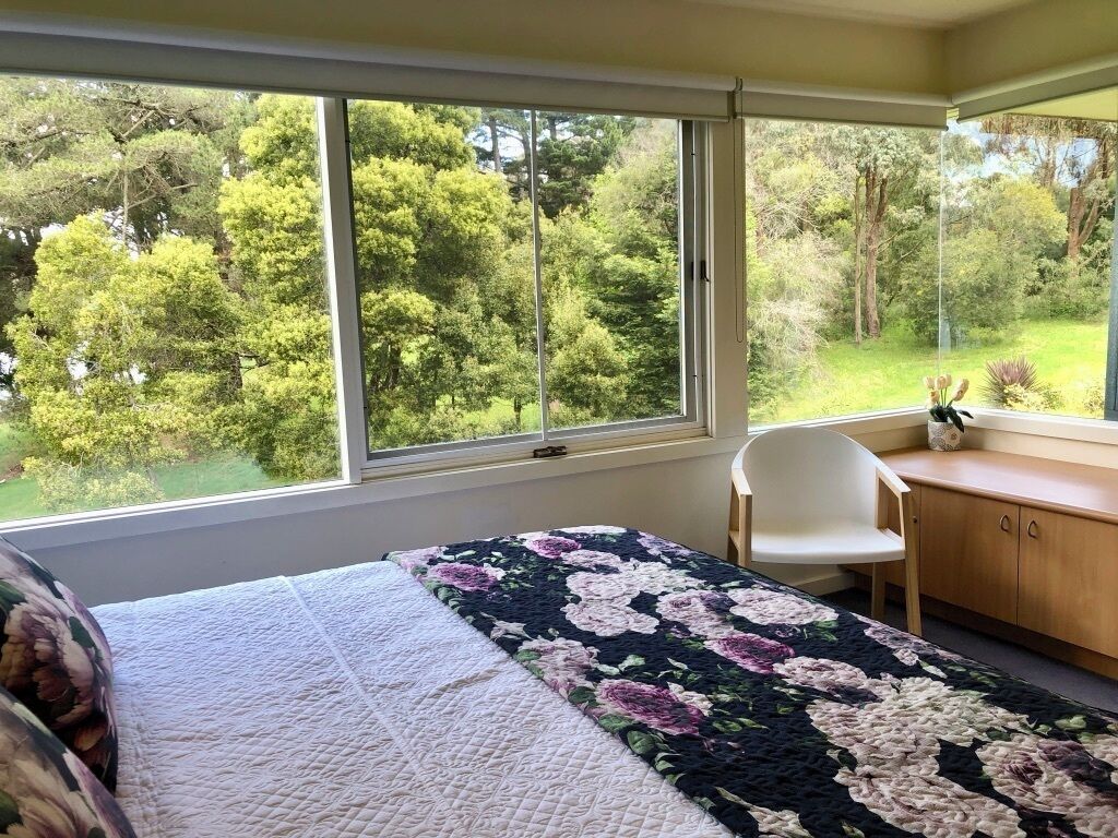 Un dormitorio con un ventanal grande, una cama con colcha estampada, una silla blanca y un escritorio de madera.