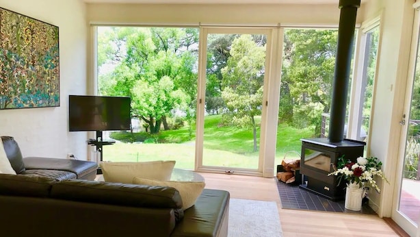 A living room with a sofa, a flat-screen TV, a fireplace, and a view of a green garden through large glass doors. - Lake Daylesford Villa 2~ lake frontage large villa
 (Daylesford)