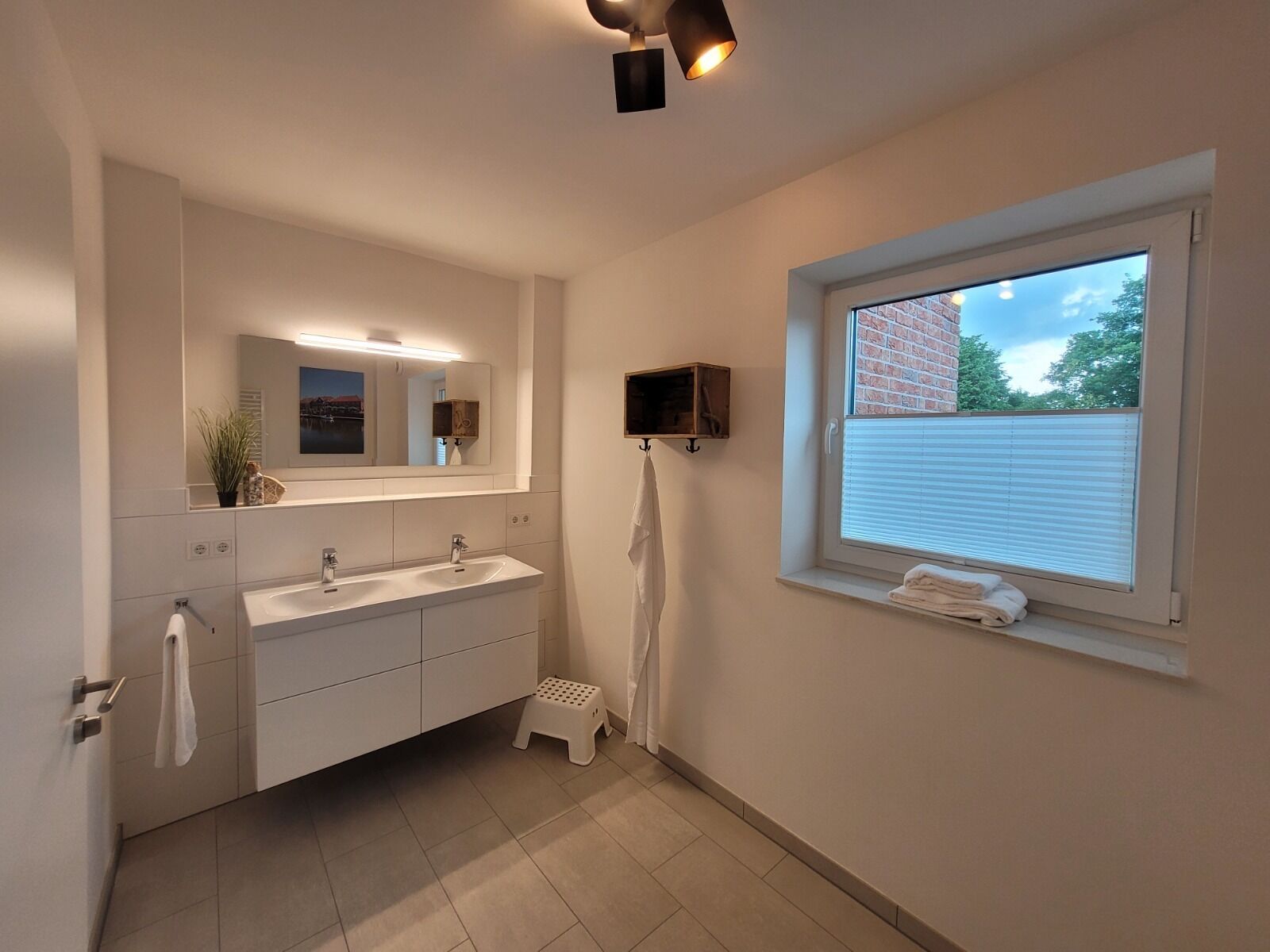Ein modernes Badezimmer mit einem großen Spiegel, zwei Waschbecken, einem Handtuchhalter und einem Fenster mit Blick auf ein Backsteinhaus.