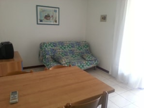 A room with a wooden table, a patterned sofa, a framed picture on the wall, and a white curtain. - Apartment With Shared Pool for 4 Guests (San Michele al Tagliamento)