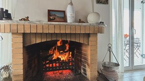 Une cheminée avec un feu qui crépite, un tableau encadré au-dessus et une bouteille de vin posée sur la manteau de la cheminée.