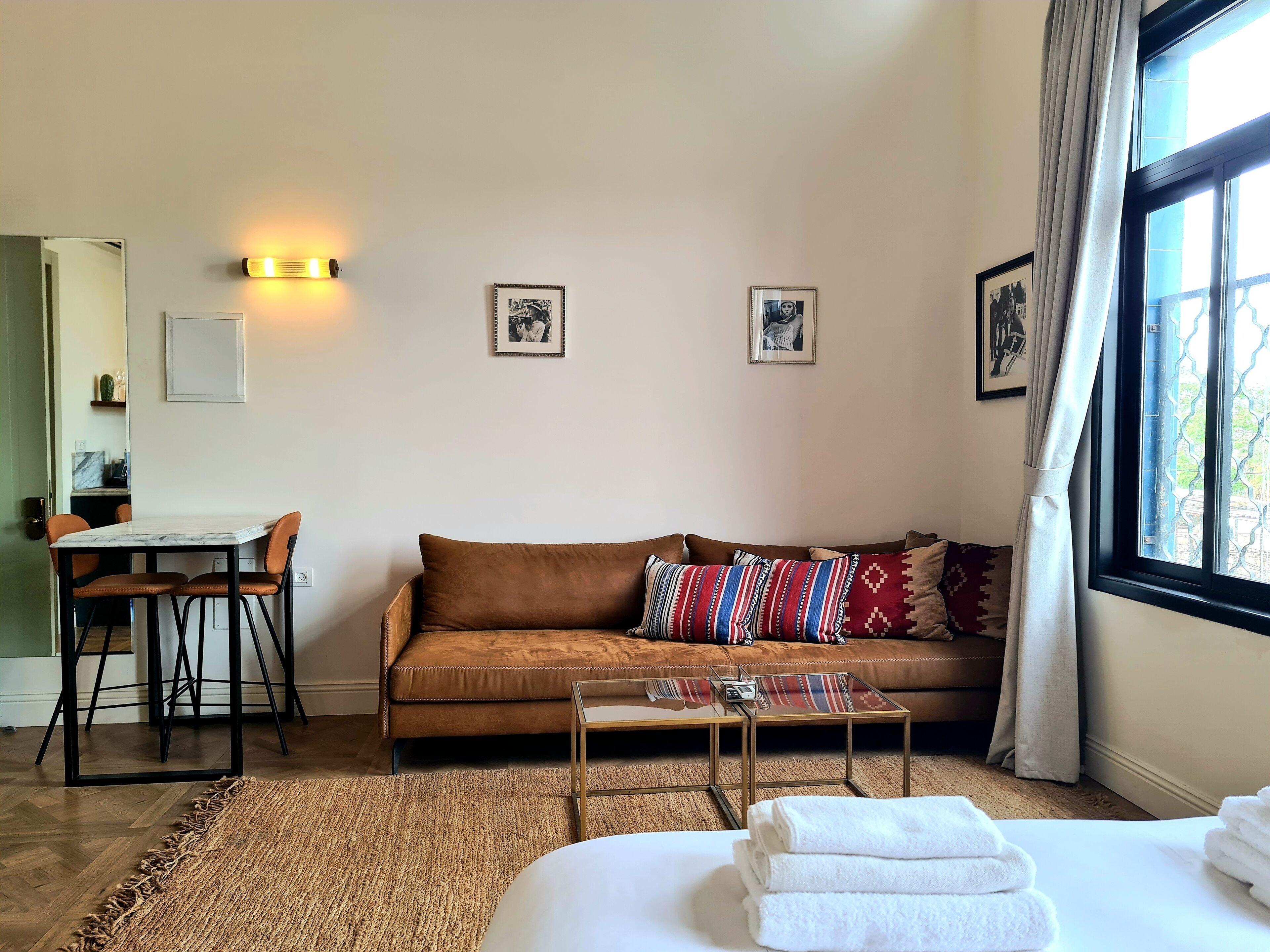 A hotel room with a brown leather sofa, a glass coffee table, a bed with white linens, a small dining table with chairs, and framed pictures on the wall.