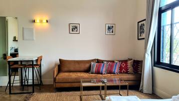 A hotel room with a brown leather sofa, a glass coffee table, a bed with white linens, a small dining table with chairs, and framed pictures on the wall.