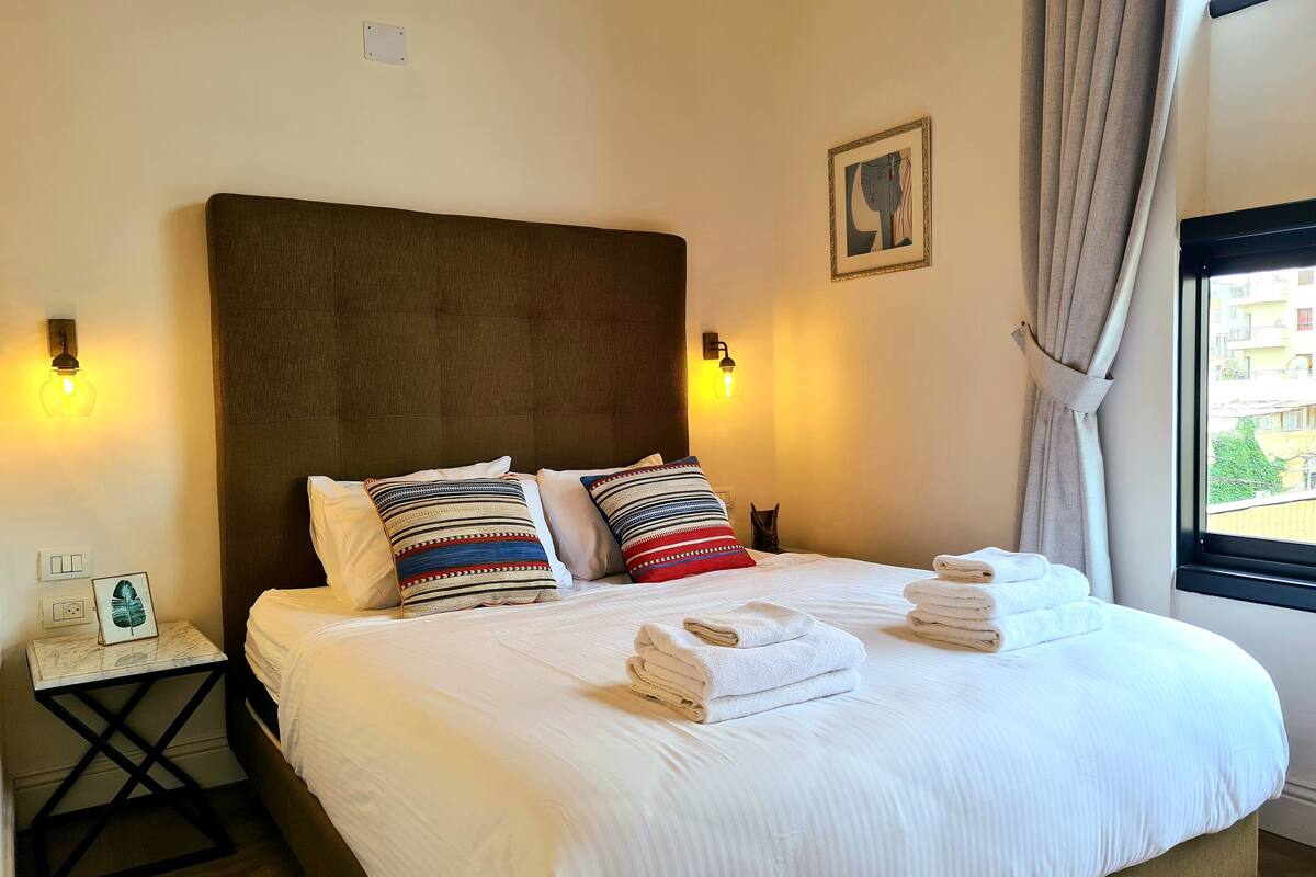 A neatly made bed with white linens and decorative pillows, a bedside table with a framed picture, and a window with curtains.