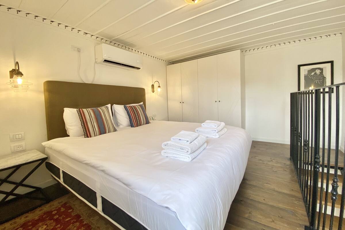 A hotel room with a large bed, a headboard, a wall-mounted air conditioner, a bedside table, and a framed picture on the wall.