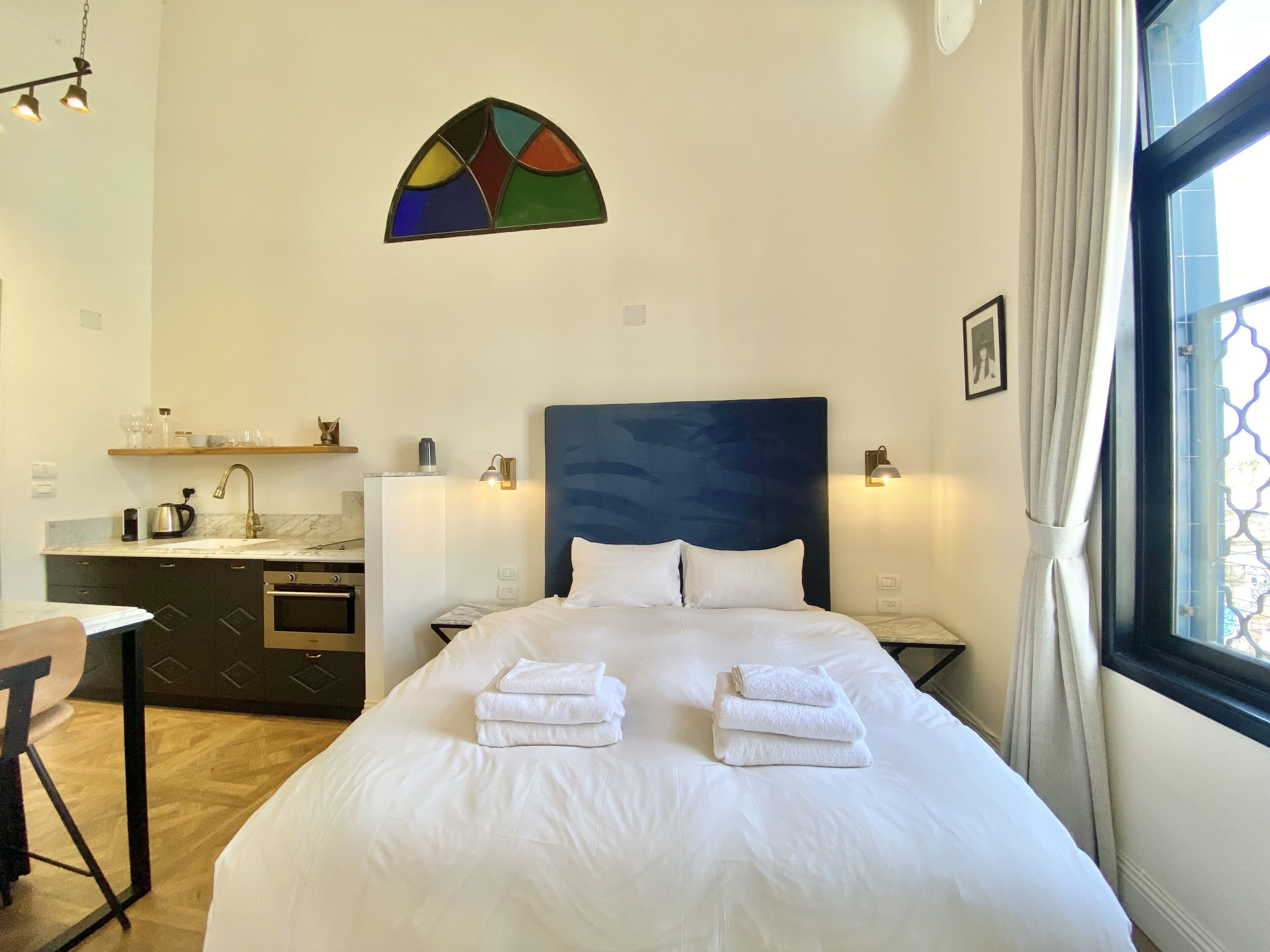 A hotel room with a bed, bedside tables, a desk, a kitchen area, and a window with curtains.