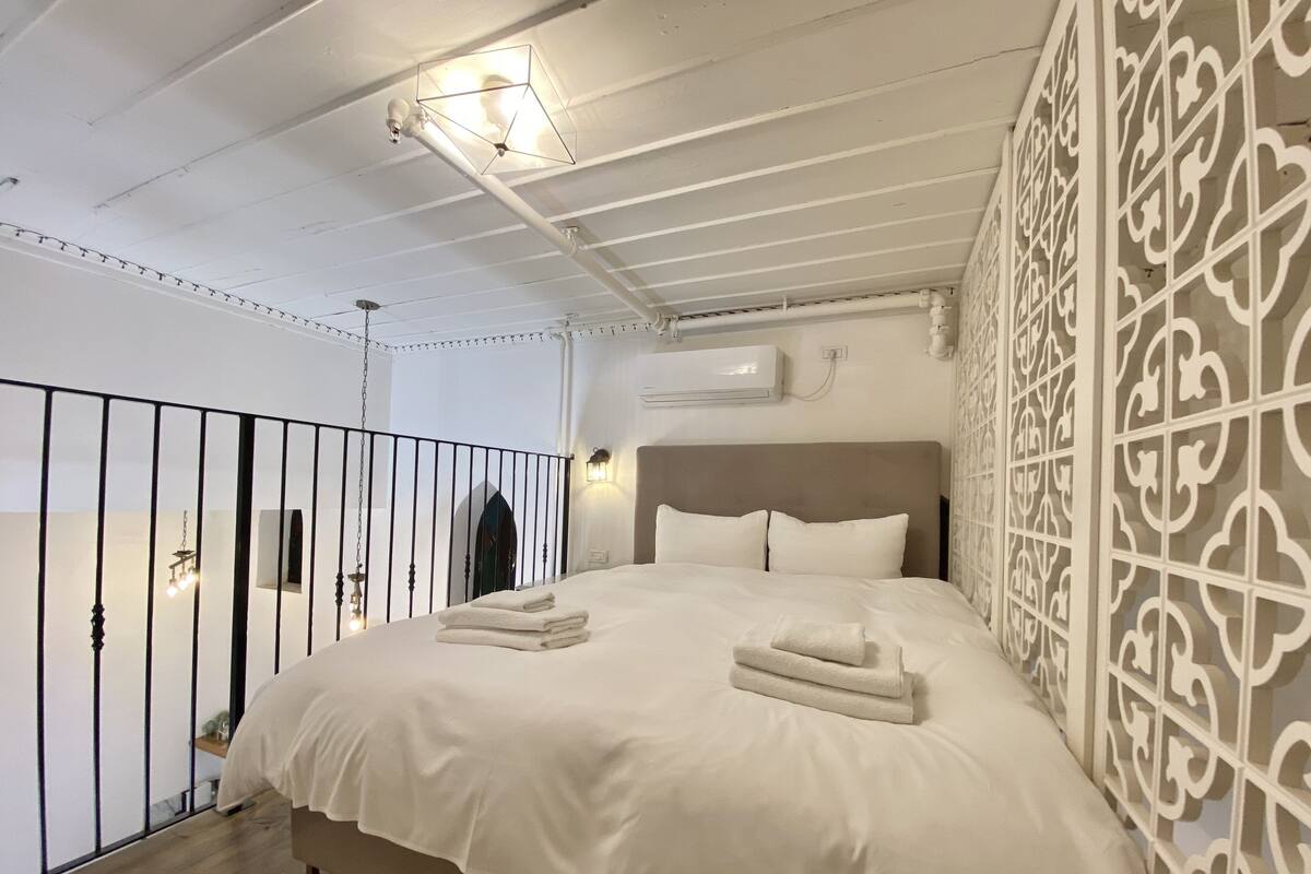 A bedroom with a bed, white linens, a headboard, a decorative wall panel, and a balcony with a railing.