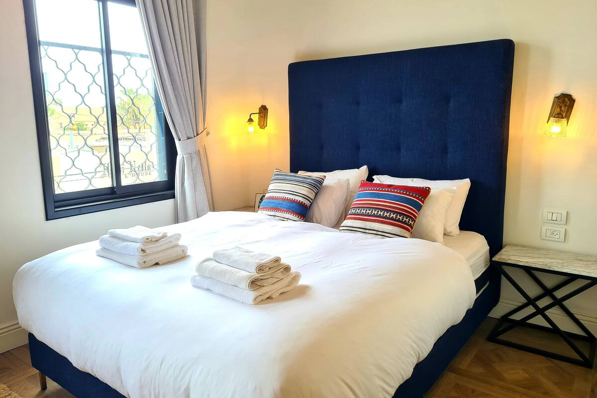 A neatly made bed with white linens and folded towels, a dark blue headboard, and two wall-mounted light fixtures.