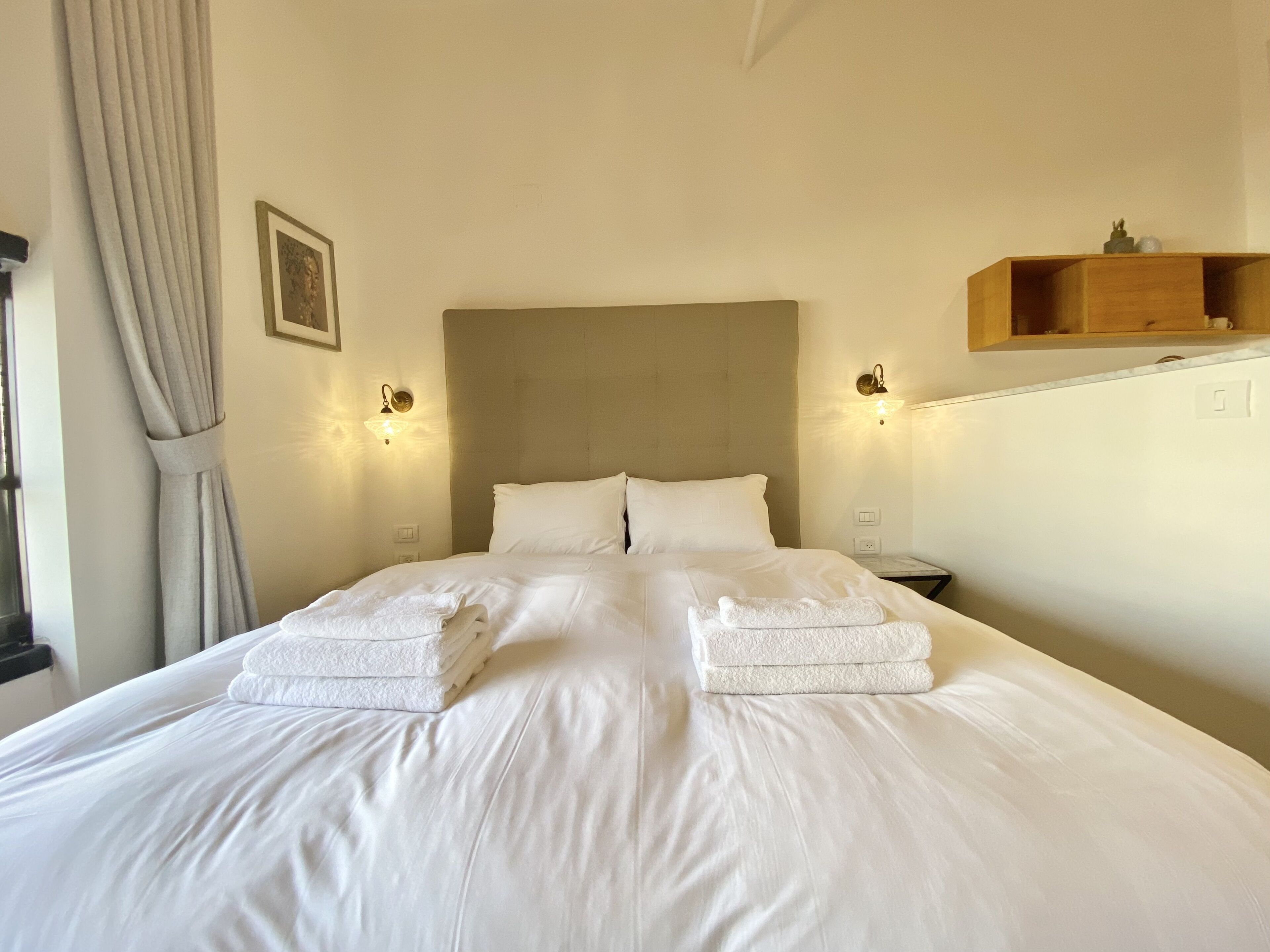Een netjes opgemaakt bed met twee opgevouwen handdoeken, een hoofdbord, nachtlampjes en een klein houten plankje boven het bed.