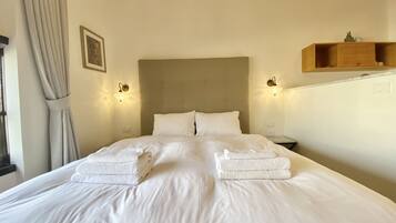 Een netjes opgemaakt bed met twee opgevouwen handdoeken, een hoofdbord, nachtlampjes en een klein houten plankje boven het bed.