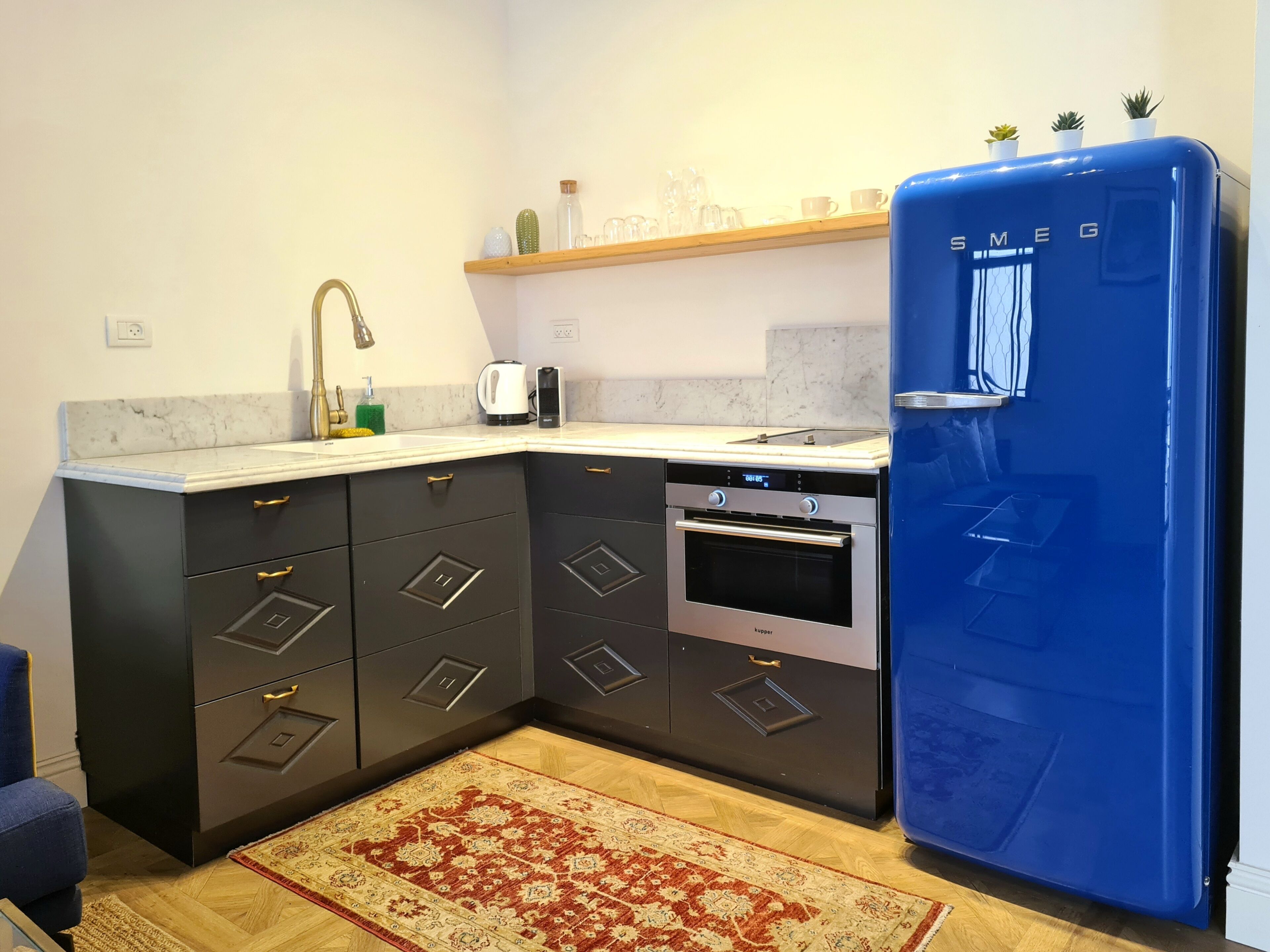 A compact kitchen with dark cabinetry, a built-in oven, a blue refrigerator, and a wooden shelf above the sink.